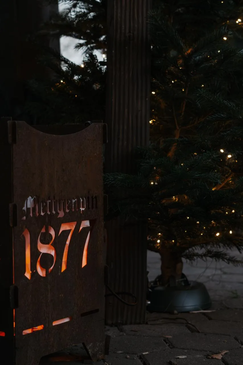 Von innen beleuchtetes Schild mit dem Schriftzug Mettgenpin 1877 vor einem Tannenbaum.