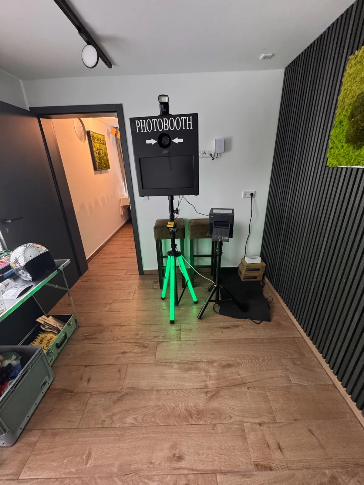 Fotobox mit der Aufschrift Photobooth in einem Flur, davor ein grün beleuchtetes Stativ.