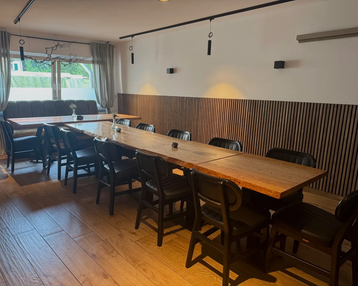 Langer Holztisch mit dunklen Stühlen vor einer Wand aus Holzlamellen, links Fenster.