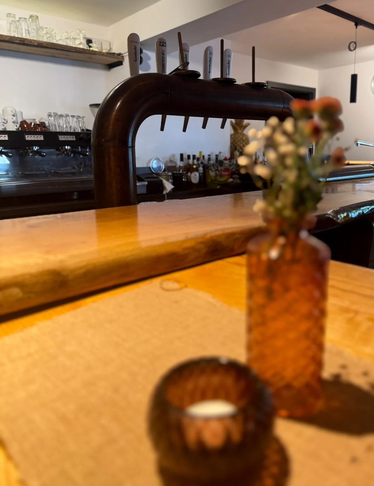 Holztheke mit Zapfhähnen im Hintergrund, im Vordergrund eine Glasvase mit Trockenblumen und ein Teelicht.
