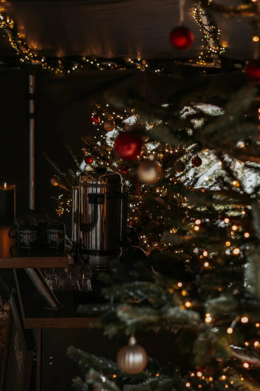Geschmückter Tannenbaum mit Lichterketten und Kugeln, im Hintergrund ein Metallbehälter für Heißgetränke.