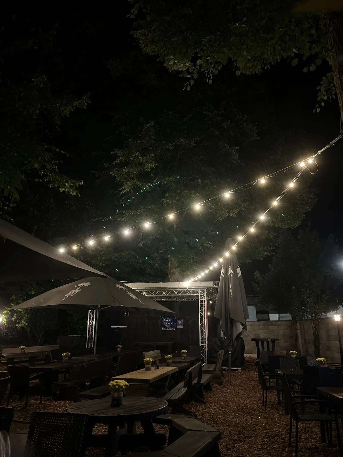 Lichterketten über dem Biergarten bei Nacht, darunter Sonnenschirme und Tische.