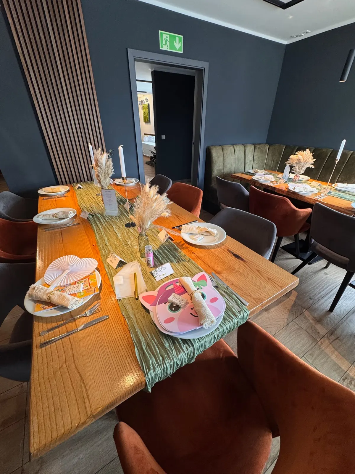 Festlich gedeckte Tafel mit grünem Tischläufer, Trockenblumen und Servietten vor einer Wand aus Holzlamellen.