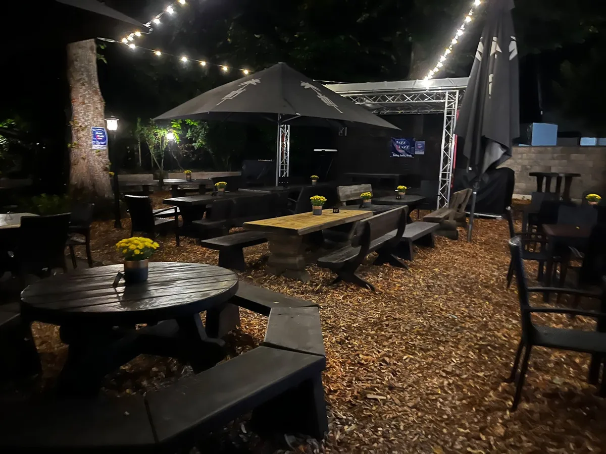 Biergarten am Abend, über Tischen und Bänken hängen Lichterketten, dazwischen Sonnenschirme.
