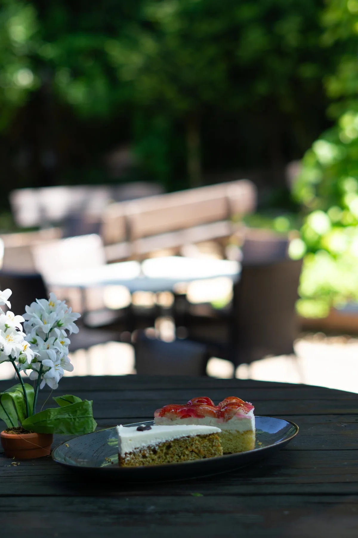 Ein Stück Torte auf einem Teller auf einem dunklen Gartentisch, im Hintergrund der Biergarten und weiße Blumen.