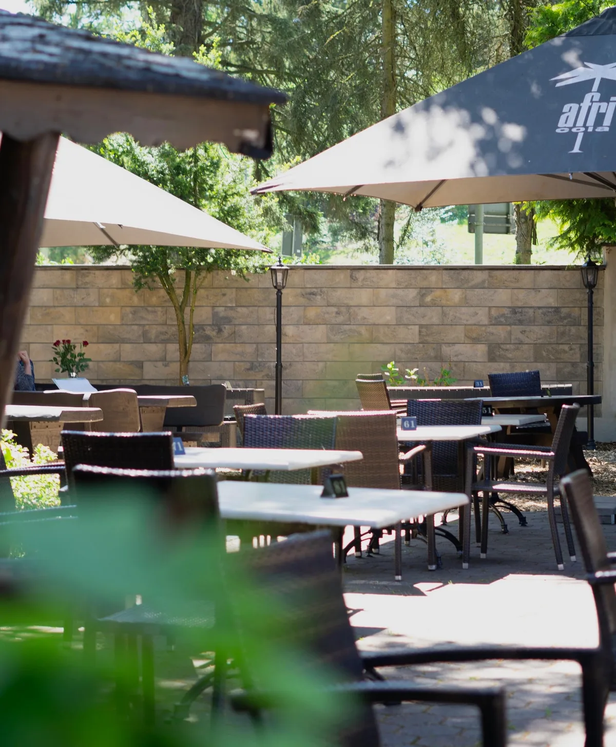 Biergarten bei Tag mit Sonnenschirmen, Tischen und Stühlen vor einer Natursteinmauer und Bäumen.